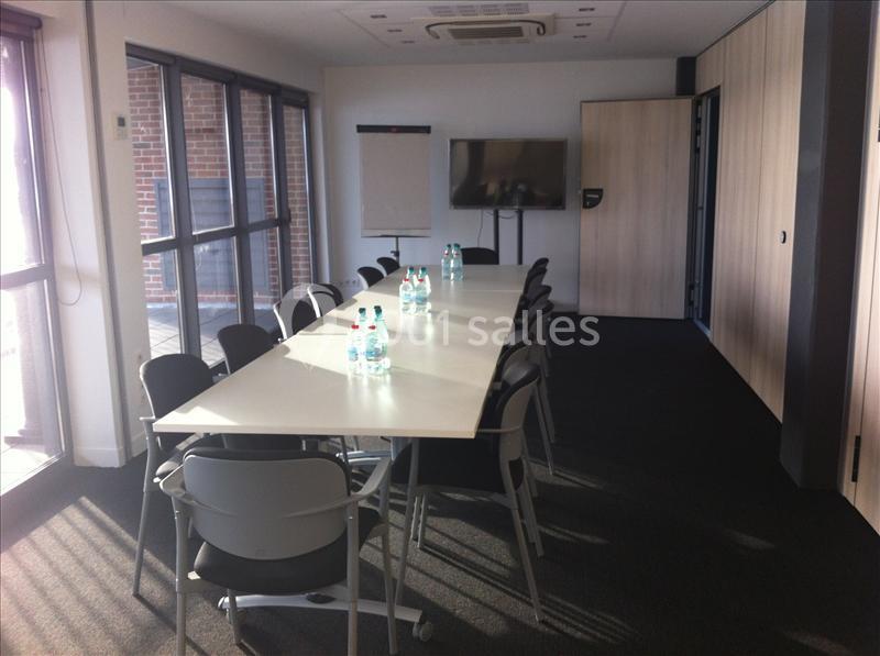Salle de réunion lumineuse avec une grande table, des chaises, un écran, un paperboard et des bouteilles d'eau.
