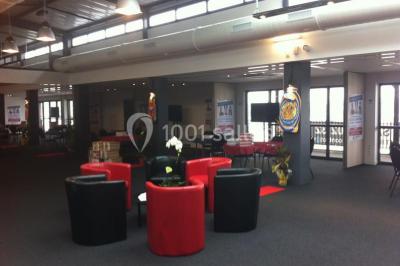 Espace intérieur lumineux avec fauteuils rouges et noirs, tables hautes, plantes et stands d'exposition en arrière-plan.