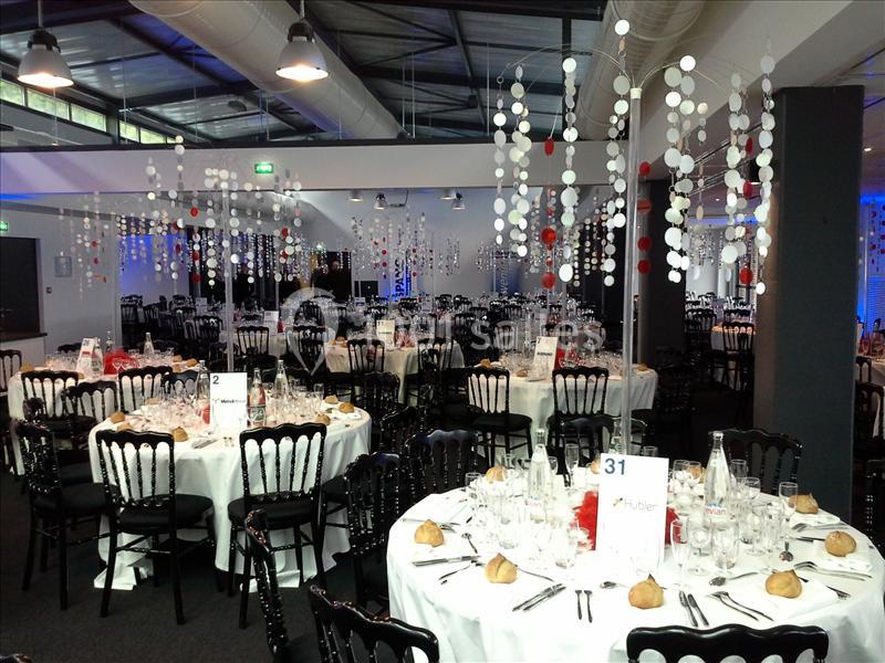 Salle de réception décorée avec des tables rondes dressées, chaises noires et suspensions blanches et rouges.