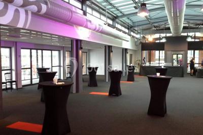 Espace intérieur lumineux avec fauteuils rouges et noirs, tables hautes, plantes et stands d'exposition en arrière-plan.