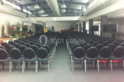 Espace intérieur lumineux avec fauteuils rouges et noirs, tables hautes, plantes et stands d'exposition en arrière-plan.