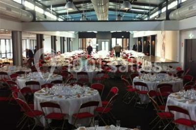 Espace intérieur lumineux avec fauteuils rouges et noirs, tables hautes, plantes et stands d'exposition en arrière-plan.