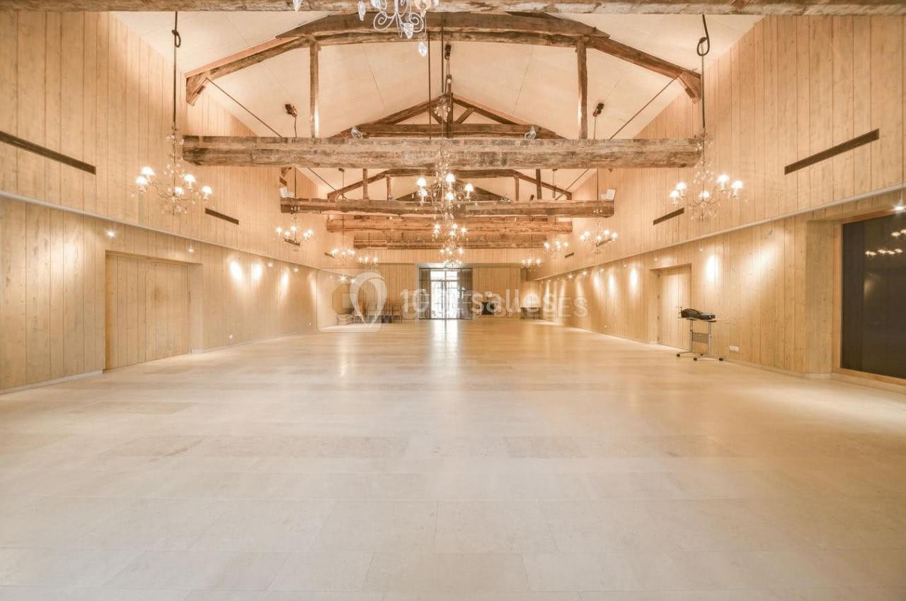 Grande salle lumineuse avec poutres apparentes, murs en bois clair, lustres suspendus et sol en pierre beige.