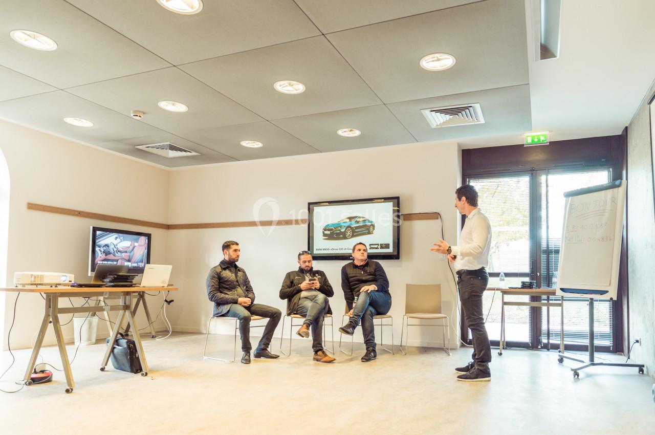 Un homme debout présente à trois personnes assises dans une salle équipée d'écrans et d'un paperboard.