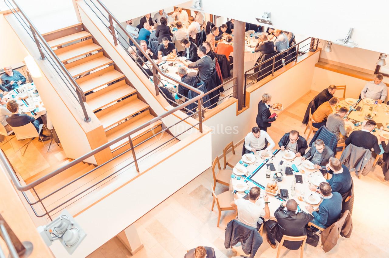 Des personnes assises autour de tables rondes lors d'un repas dans une salle lumineuse avec un escalier visible.