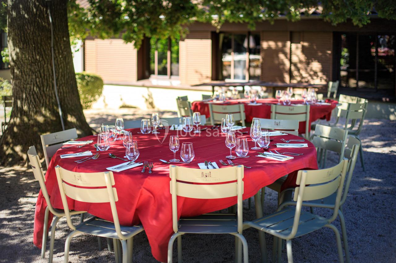 Tables rondes dressées avec nappes rouges, couverts et verres, disposées en extérieur sous des arbres.
