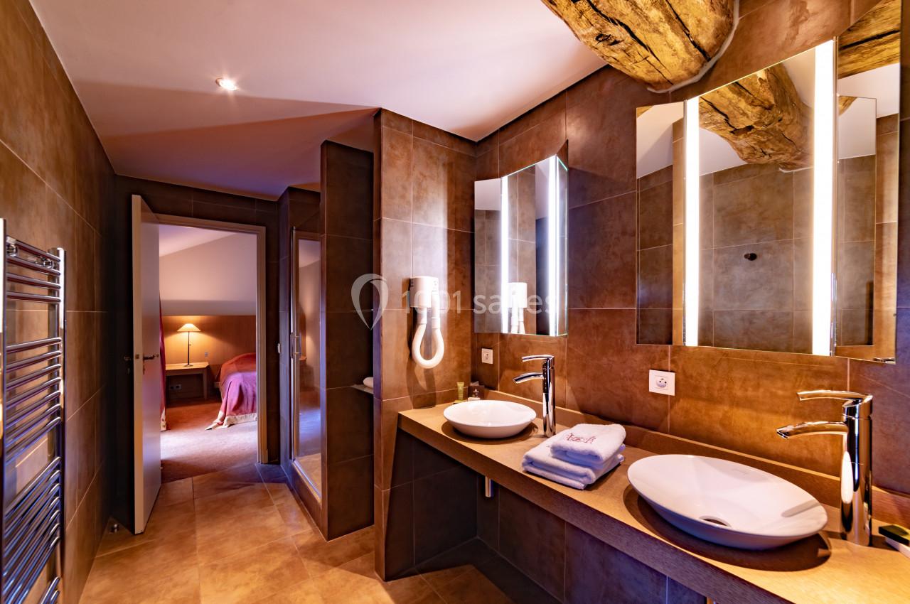 Salle de bain moderne avec deux vasques, miroirs éclairés, sèche-cheveux mural et vue sur une chambre adjacente.