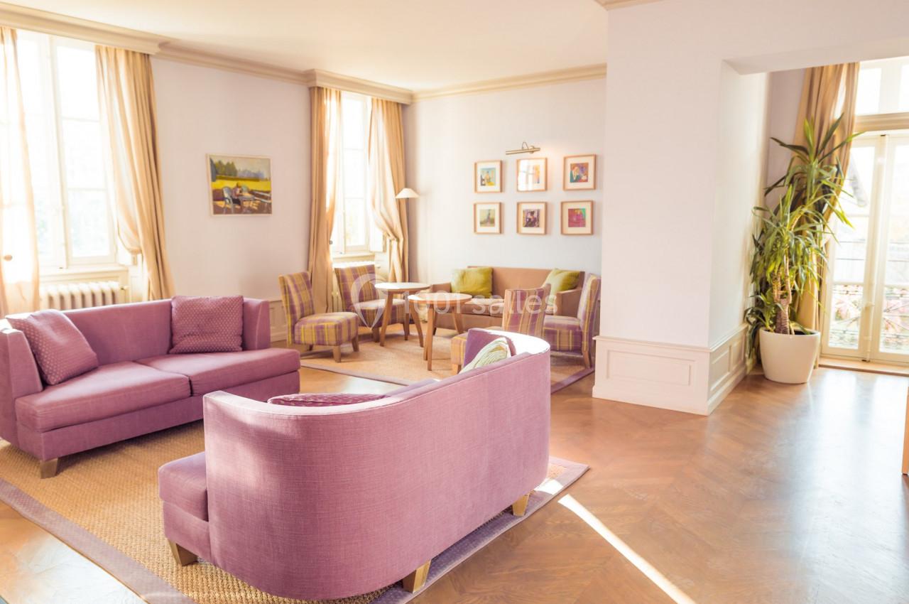 Salon lumineux avec mobilier violet, parquet en bois, grandes fenêtres, plantes et tableaux décoratifs au mur.