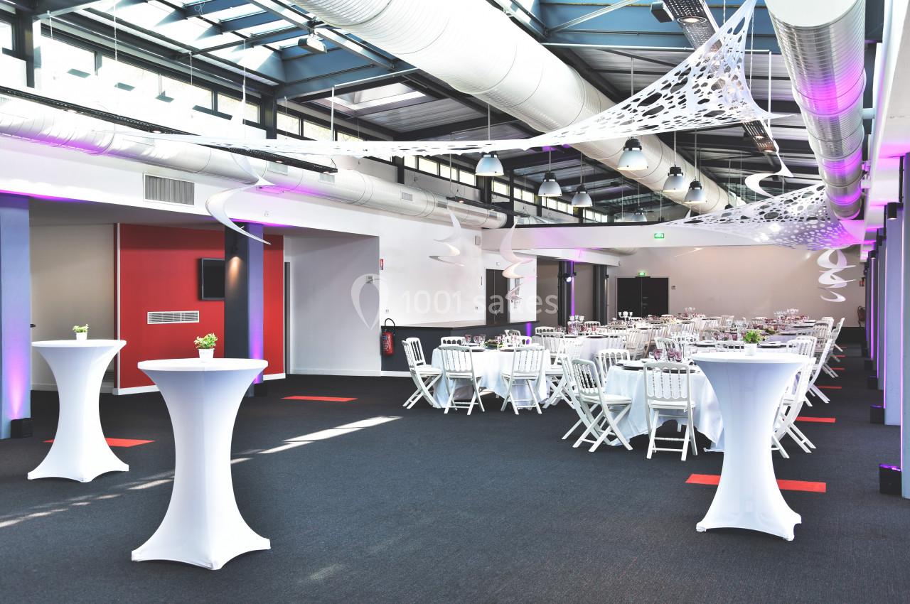 Salle de réception moderne avec tables rondes dressées, chaises blanches et décorations suspendues.