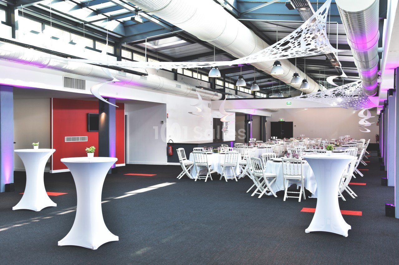 Salle de réception moderne avec tables rondes dressées, chaises blanches et décorations suspendues.