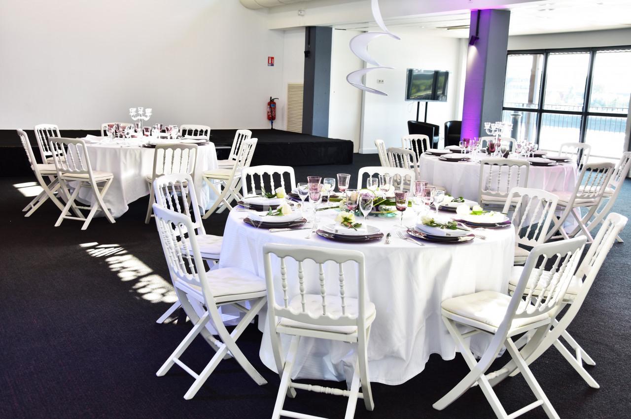 Salle lumineuse avec tables rondes dressées, nappes blanches, chaises pliantes blanches et décoration élégante.