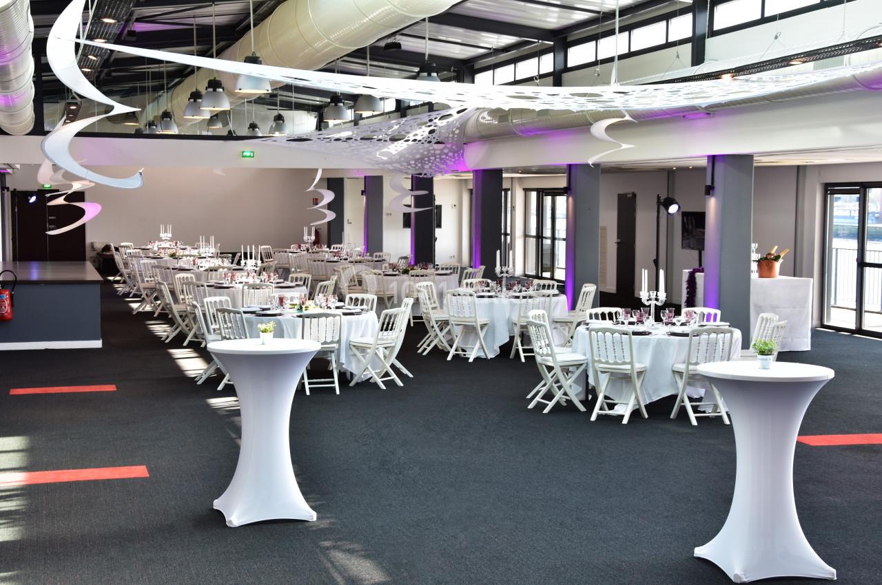 Salle de réception lumineuse avec tables rondes dressées, chaises blanches et décorations suspendues modernes.