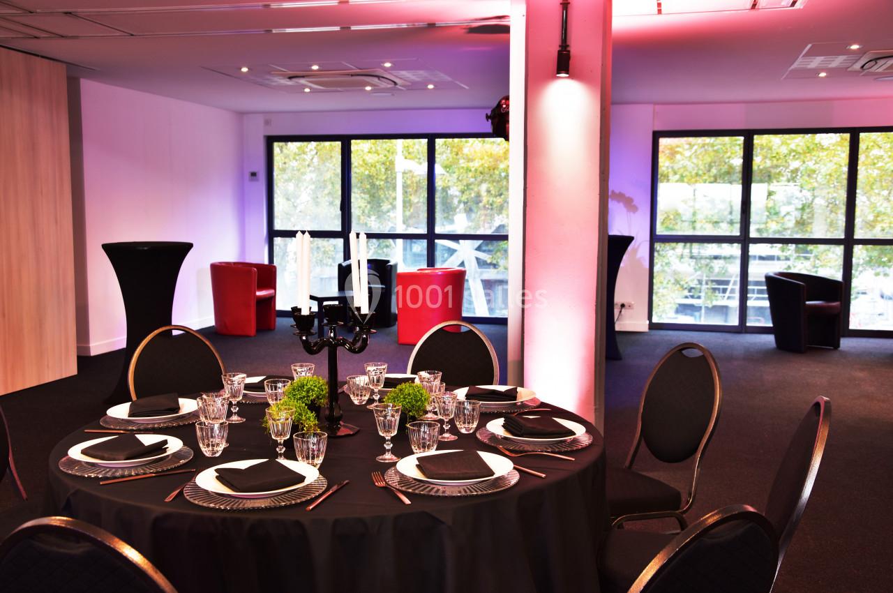 Salle de réception moderne avec une table ronde dressée, chandeliers noirs, et grandes baies vitrées donnant sur des arbres.