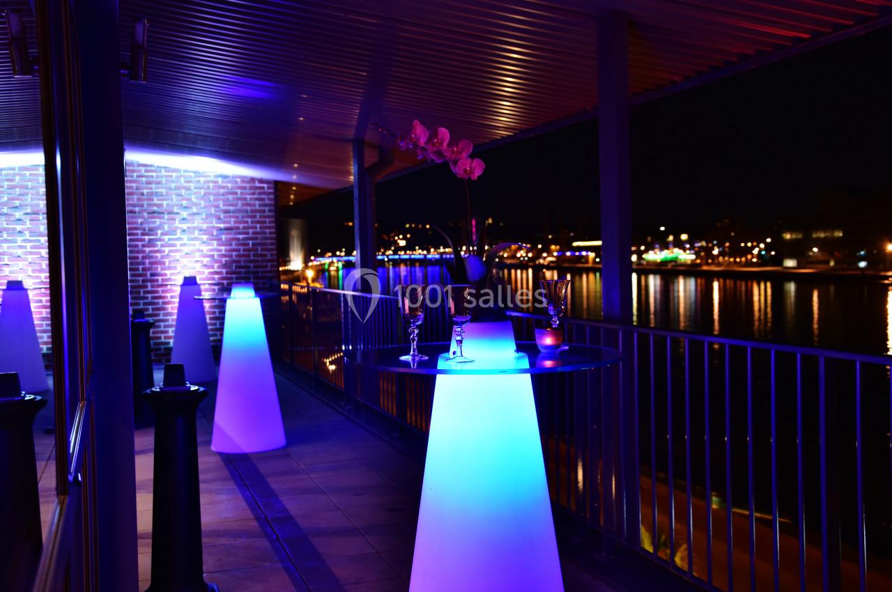 Terrasse éclairée de nuit avec des tables lumineuses, une orchidée et une vue sur des lumières reflétées dans l'eau.