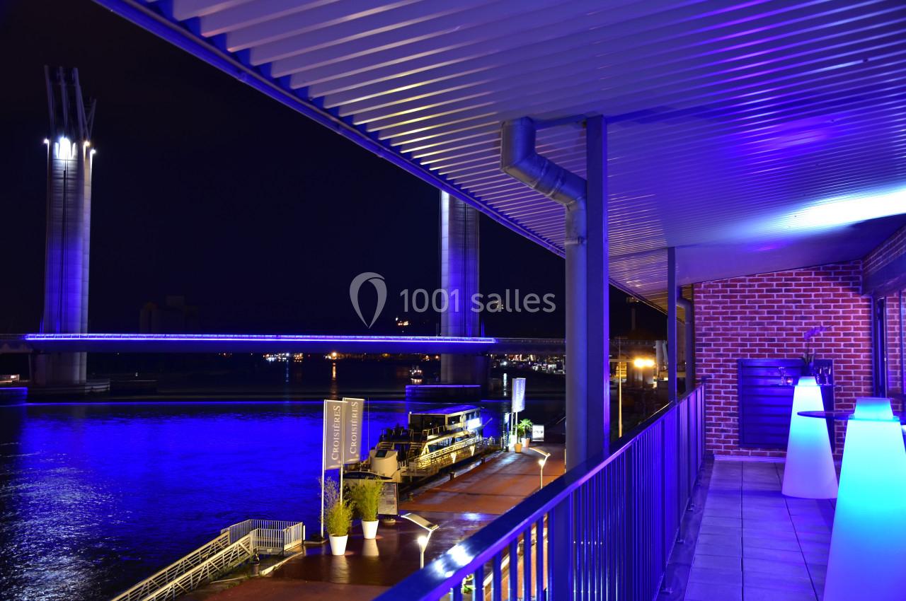 Vue nocturne d'une terrasse éclairée en bleu avec une rivière et un pont illuminé en arrière-plan.