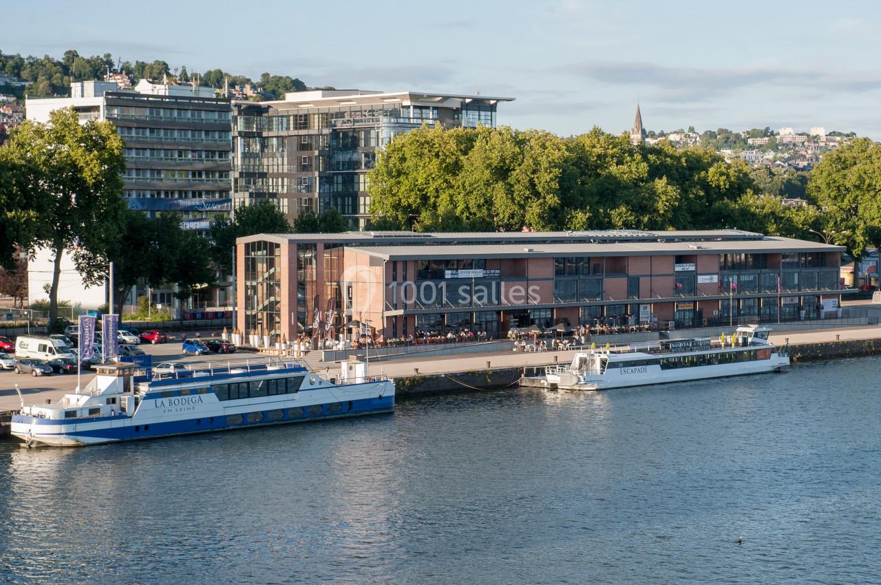 Vue d'un quai avec deux bateaux amarrés devant un bâtiment moderne entouré d'arbres et d'immeubles.