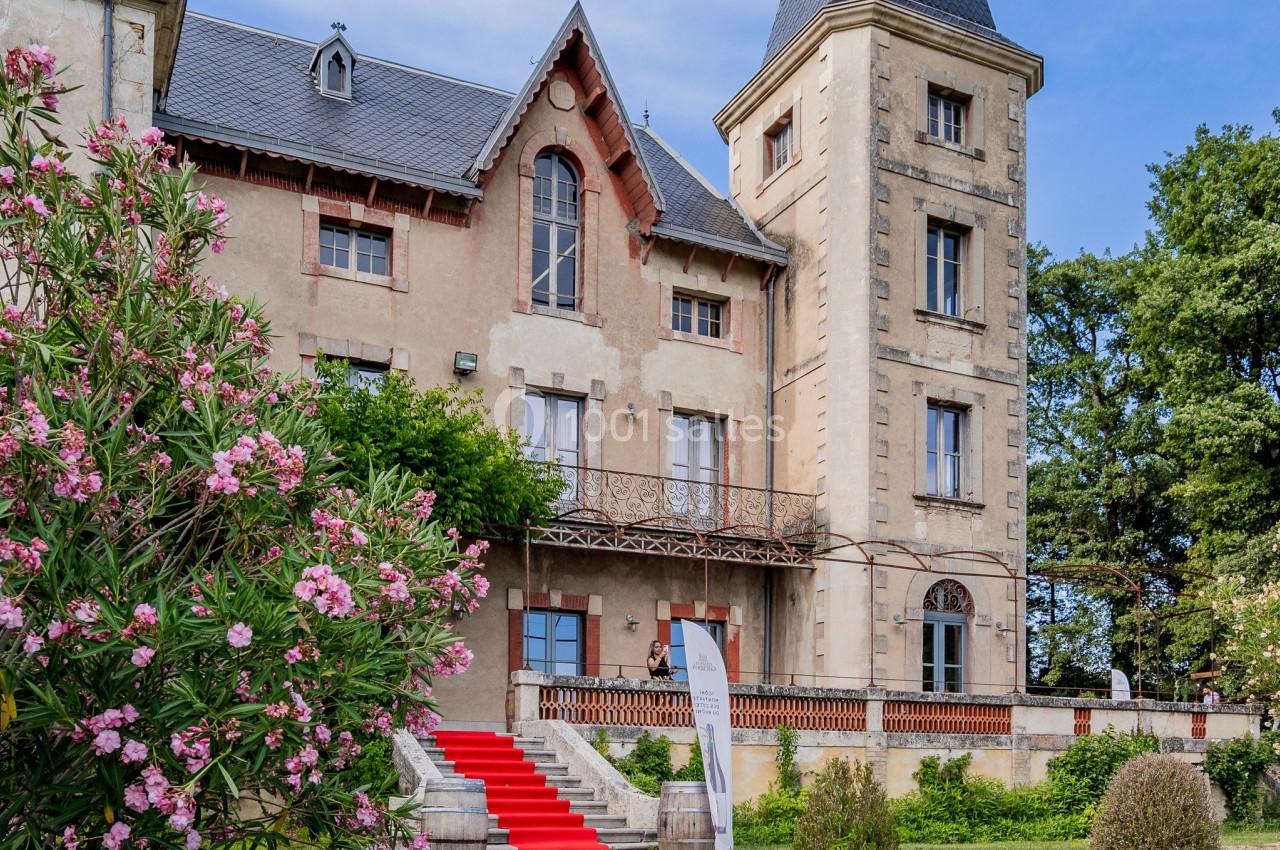 Façade d'un manoir avec une tour, escalier rouge, jardin fleuri et ciel dégagé en arrière-plan.