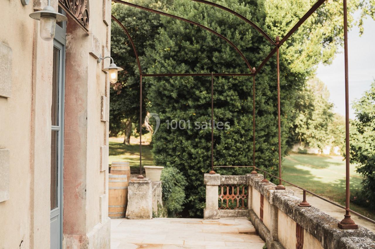 Terrasse en pierre bordée de balustrades et d'arches métalliques, donnant sur un jardin arboré.