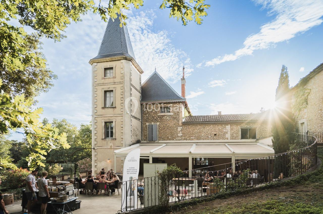 Vue d'un bâtiment en pierre avec une tour, une terrasse animée et des convives, entouré de verdure sous un ciel ensoleillé.