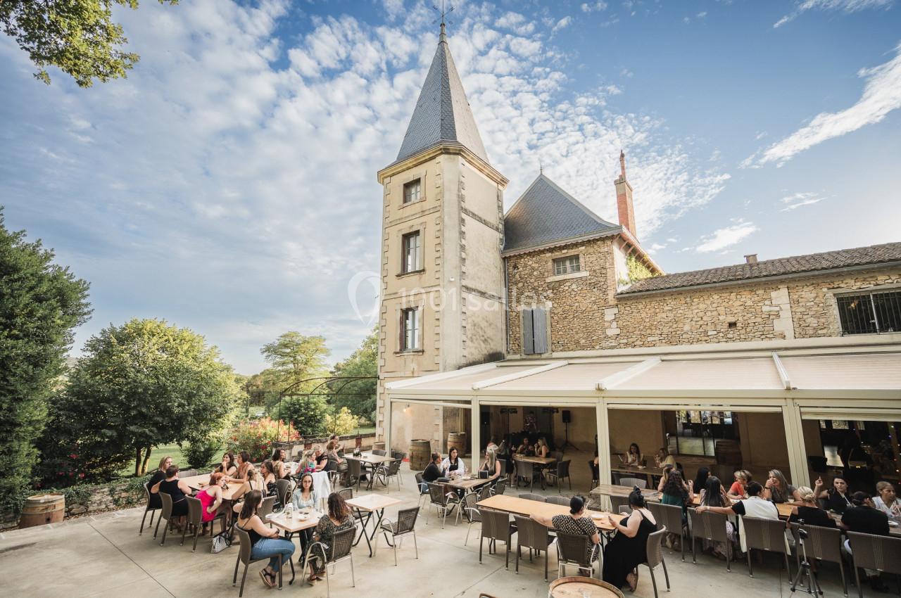 Groupe de personnes attablées en terrasse devant un bâtiment en pierre avec une tour, sous un ciel partiellement nuageux.