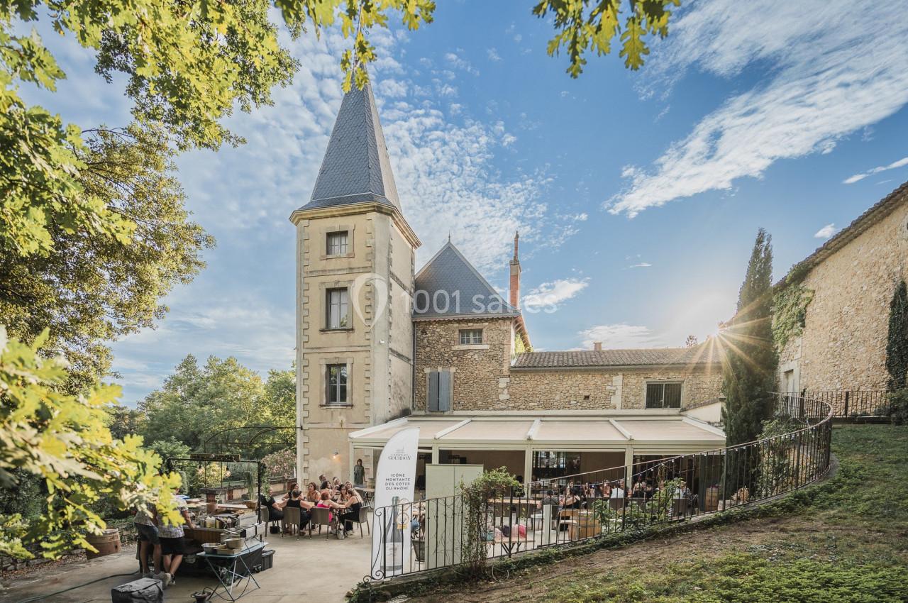 Vue d'un bâtiment en pierre avec une tour, entouré de verdure, abritant une terrasse animée par des convives.
