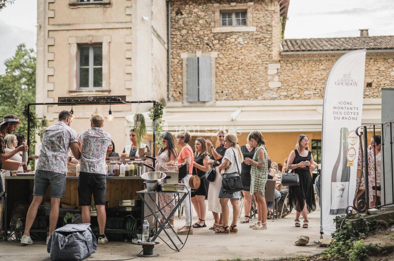 Des personnes font la queue devant un stand en plein air, près d'un bâtiment en pierre, lors d'un événement convivial.