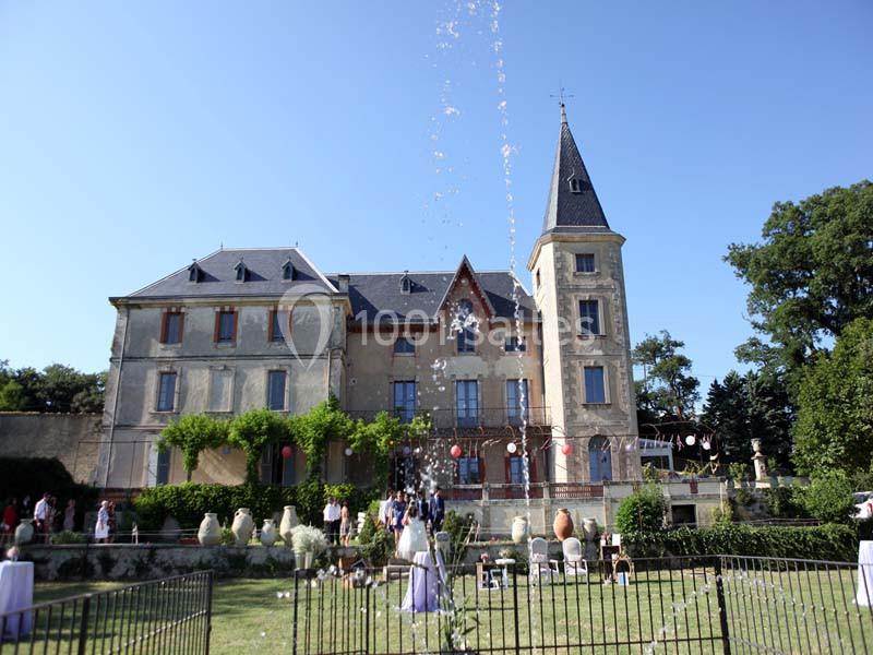 Façade d'un château entouré de verdure, avec une fontaine en premier plan et des personnes dans le jardin.
