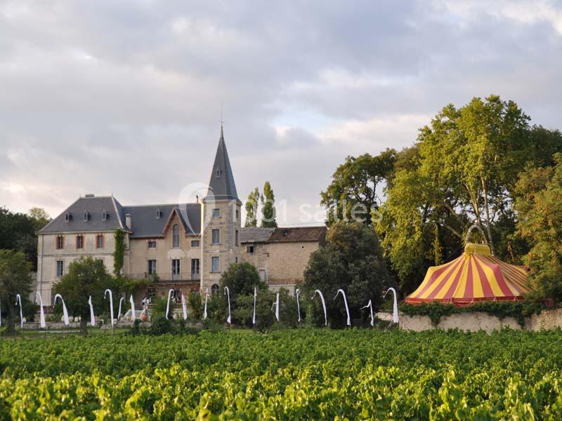 Château en pierre entouré de vignes, avec une tente de cirque jaune et rouge à proximité, sous un ciel nuageux.