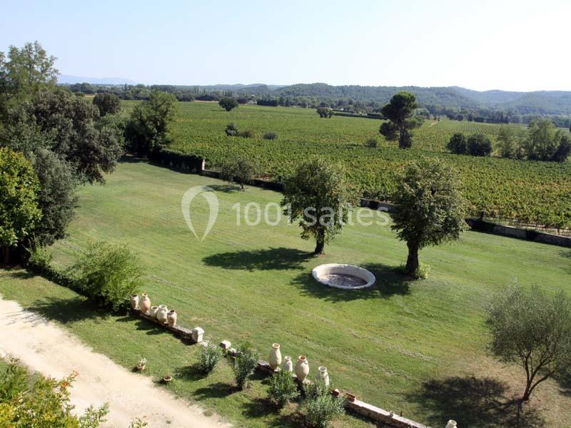 Vue d'un grand jardin verdoyant avec deux arbres, une fontaine centrale et des champs en arrière-plan.