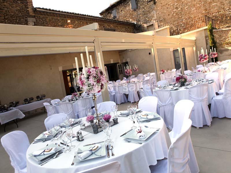 Salle de réception extérieure avec tables rondes décorées de nappes blanches, fleurs et chandeliers, entourée de murs en…