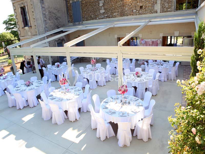 Tables rondes décorées pour un événement en extérieur sous une structure ouverte, devant un bâtiment en pierre.