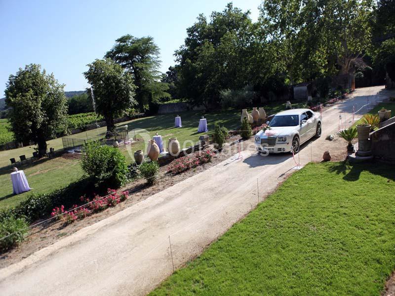 Voiture blanche roulant sur une allée gravillonnée bordée de pelouses, pots décoratifs et arbres en arrière-plan.