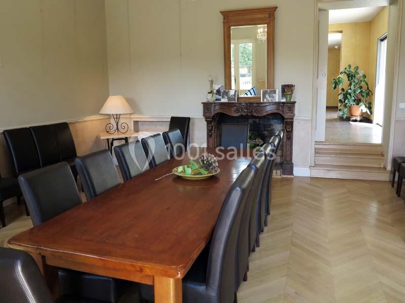 Salle à manger avec une grande table en bois entourée de chaises noires, cheminée en arrière-plan et lumière naturelle.