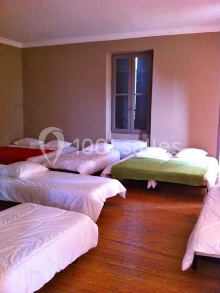 Dortoir avec plusieurs lits simples alignés, draps blancs et quelques couvre-lits colorés, sur un sol en parquet.