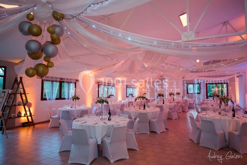 Salle de réception décorée avec des tables rondes, nappes blanches, guirlandes lumineuses et suspensions dorées.