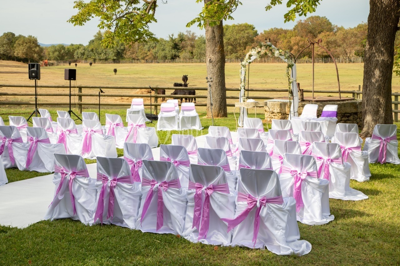 Chaises blanches décorées de rubans roses disposées en extérieur pour une cérémonie, avec une arche fleurie en arrière-plan.