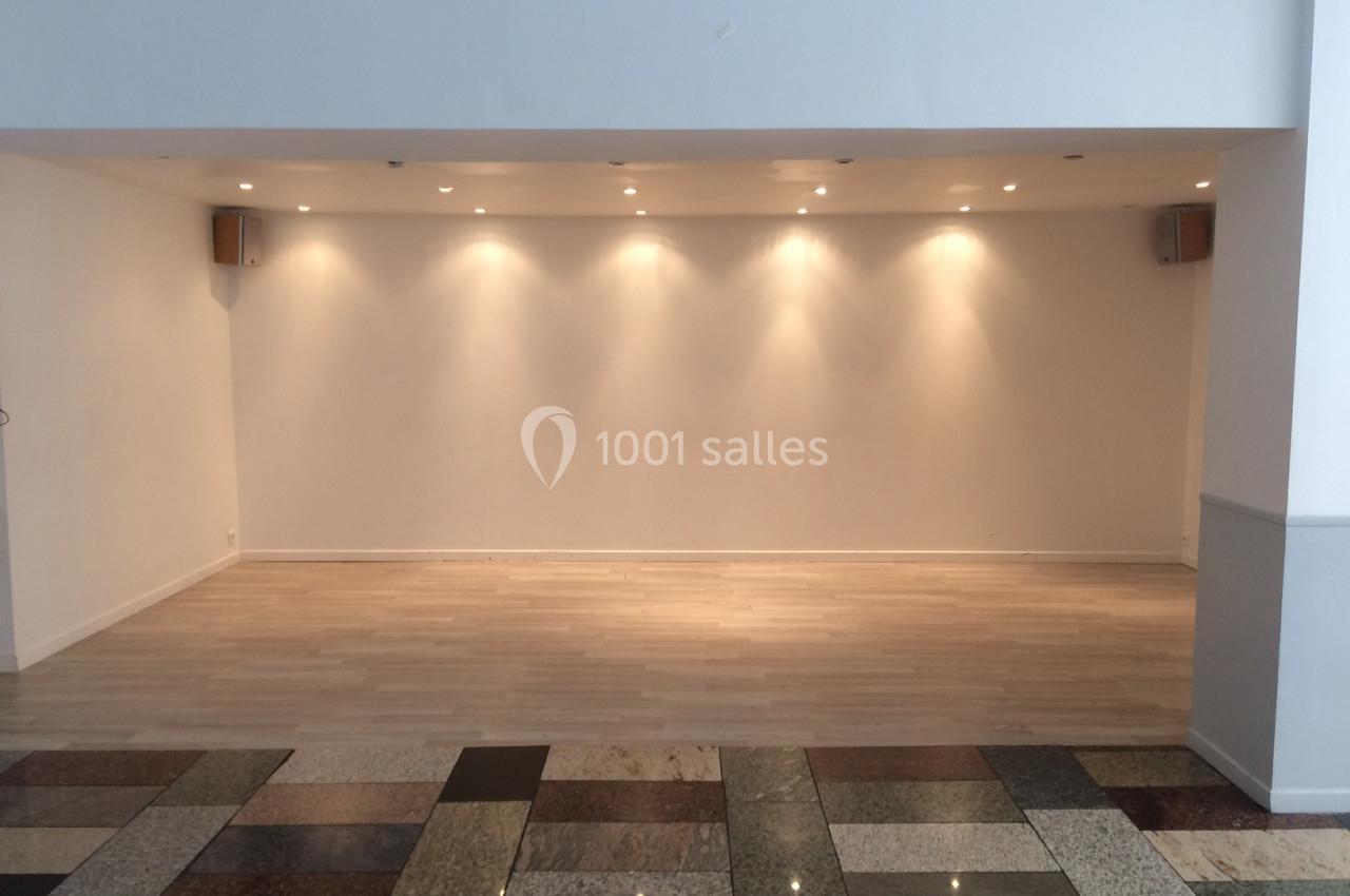 Salle vide avec un mur blanc éclairé par des spots au plafond, parquet clair et sol en carrelage multicolore au premier plan.