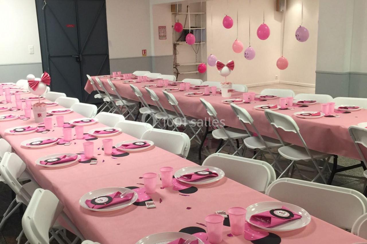 Salle décorée pour une fête avec des tables couvertes de nappes roses, vaisselle assortie et suspensions décoratives.