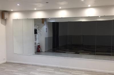 Salle lumineuse avec un grand miroir mural, sol en parquet clair et murs blancs, éclairée par des spots au plafond.