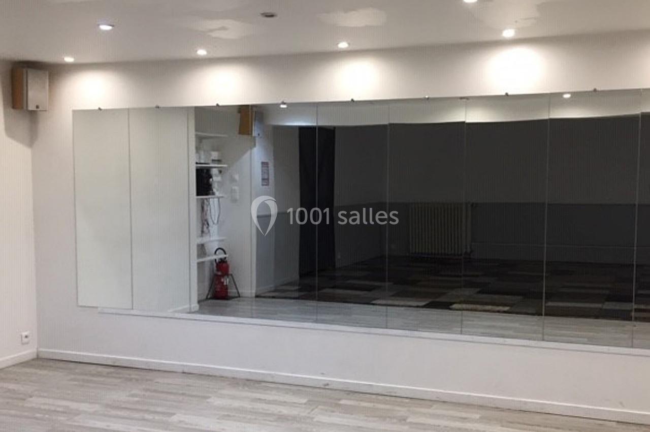 Salle lumineuse avec un grand miroir mural, sol en parquet clair et murs blancs, éclairée par des spots au plafond.