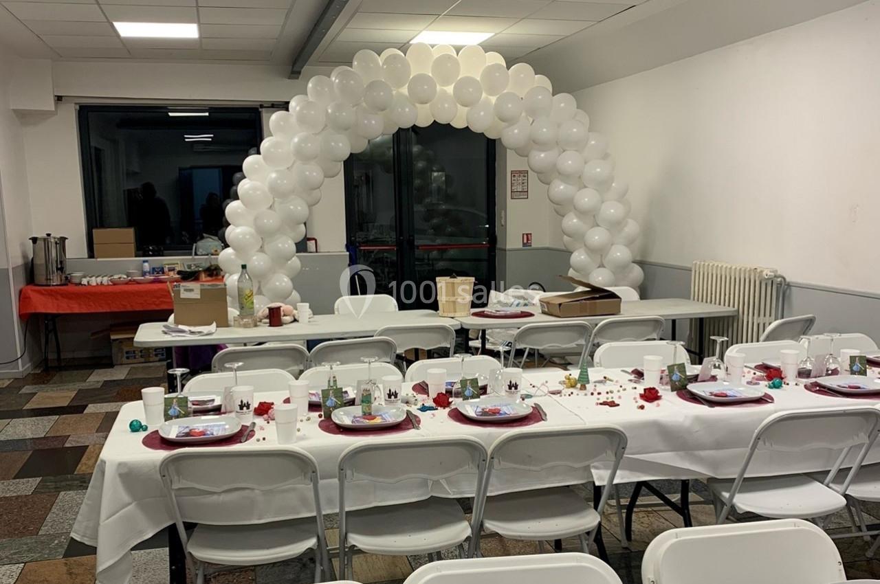 Salle décorée pour un événement avec tables dressées, nappes blanches, et arche de ballons blancs en arrière-plan.