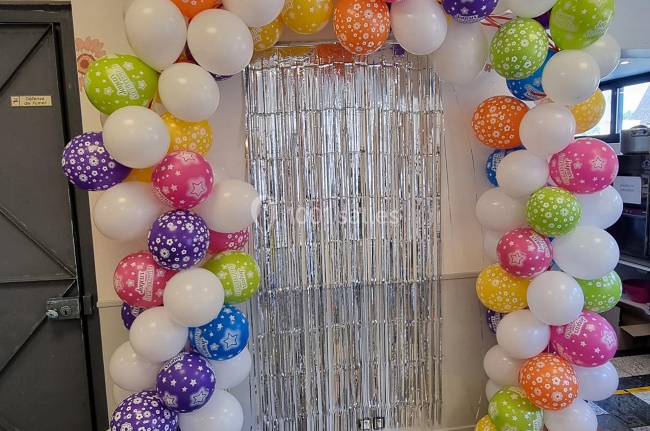 Arche décorative en ballons colorés entourant un rideau métallique argenté dans un espace intérieur.