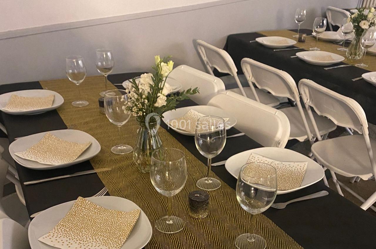 Table dressée avec nappes noires et dorées, vaisselle blanche, verres à pied et petits bouquets de fleurs blanches.
