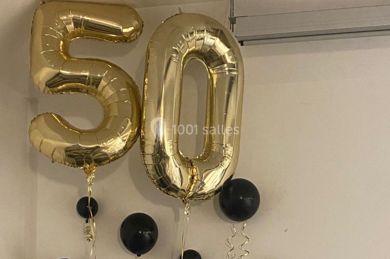 Ballons dorés formant le chiffre 50 entourés de ballons noirs suspendus dans une pièce blanche.