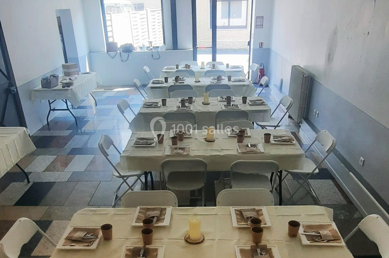 Salle aménagée avec des tables rectangulaires dressées pour un repas, chaises blanches pliantes et lumière naturelle.