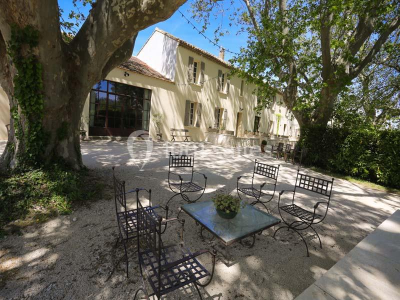 Cour ombragée avec des tables et chaises en fer forgé devant une maison en pierre claire entourée d'arbres.