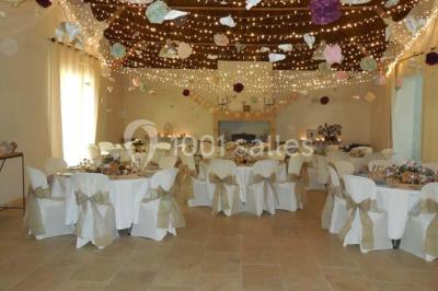 Façade d'une maison provençale avec terrasse ombragée, tables en fer forgé et grand arbre au premier plan.