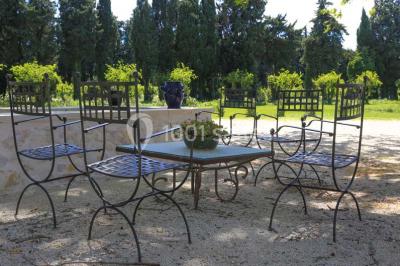 Façade d'une maison provençale avec terrasse ombragée, tables en fer forgé et grand arbre au premier plan.