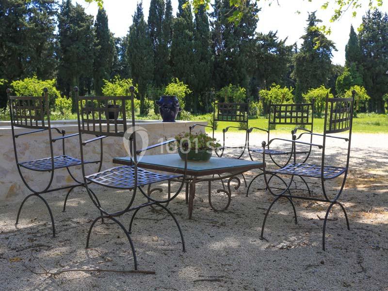 Chaises en fer forgé et table basse sur une terrasse en gravier, entourées d'arbres et de verdure.