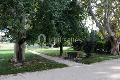 Façade d'une maison provençale avec terrasse ombragée, tables en fer forgé et grand arbre au premier plan.